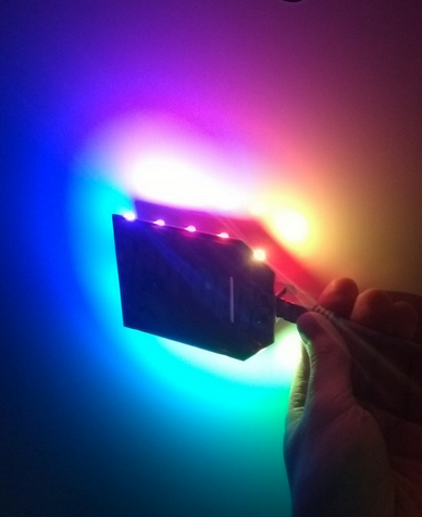 A PCB with 12 RGB leds around its edge glowing in rainbow colors in front of a white background.